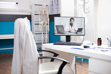 Obraz premium Caucasian doctor videocall displayed on desktop pc positioned on table of vacant hospital office. Computer screen shows healthcare expert conducting virtual medical consultation with nobody present.