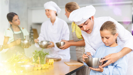 In restaurant kitchen during children cooking class, male chef help small girl thoroughly stir homogeneous liquid dough with whisk. In background, blurry participants are standing near table with food