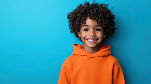 Happy young African American boy with curly hair wearing a bright orange hoodie standing against a solid blue background