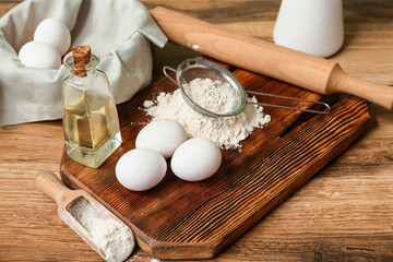 Cutting board with flour, fresh eggs, oil and utensils for preparing dough on wooden table