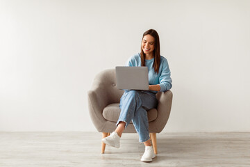 Happy Caucasian lady sitting in armchair with modern laptop, studying remotely or having business...