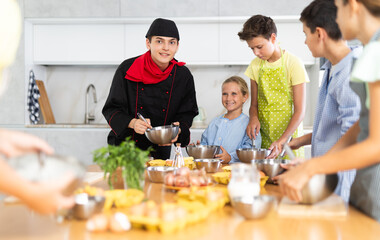 During cooking class, guy cook in black uniform stands near kitchen table with bowl in hands tells children participants culinary ticks and tricks. Man shows example of successful mobile dough