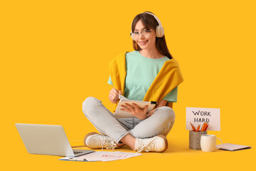 Teenage girl in headphones with papers and laptop working online on yellow background