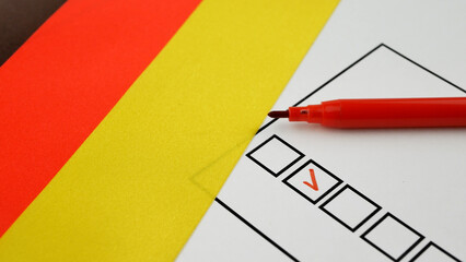 Democracy in action during German elections. Voters hand holds red marker, placing checkmark next to candidate. German flag in background. Symbolizing election integrity, civic engagement