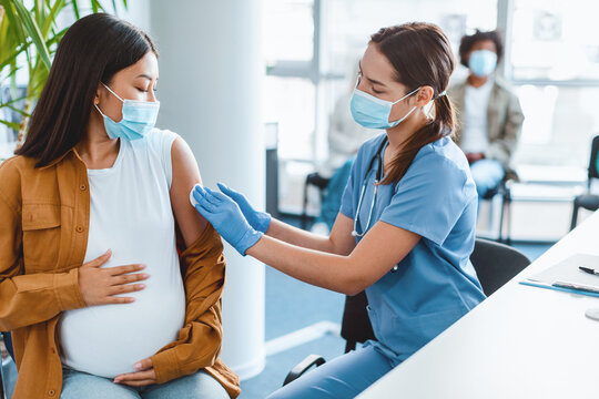 Nurse in face mask and gloves disinfecting skin of pregnant patient, rubbing shoulder with cotton pad before injection, asian lady hug belly, clinic background. flu coronavirus treatment concept