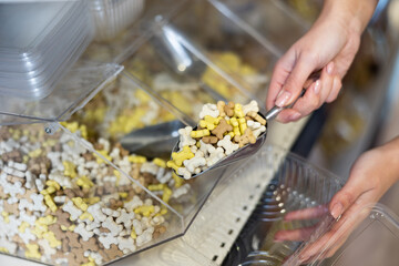 Female hands holding metal scoop with nutritious dog snacks in form of multicolored small bones, pouring into plastic container in pet store