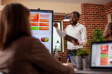 Businessman with clipboard in hand, using digital monitor to explain analytics summary in meeting. African american entrepreneur doing project presentation, planning marketing strategy with his team.