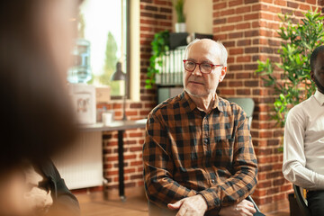 Retired white man with glasses, sitting and discussing with group at therapy session in brick wall room. Caucasian male pensioner attending rehab program and sharing his struggles with community.