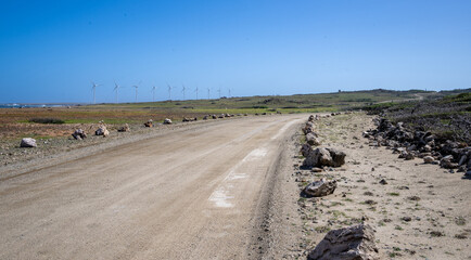 A row of giant wind turbines stands along a dusty path, set against a vibrant blue sky. The barren landscape highlights renewable energy's potential in harnessing wind power in remote areas.