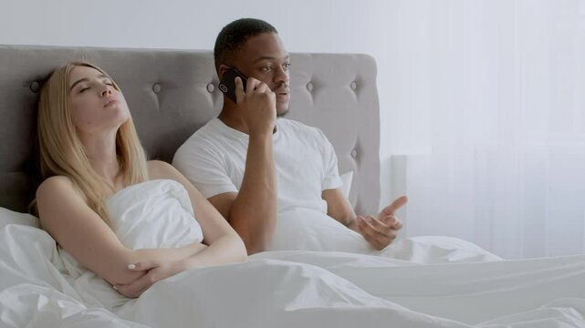 In a cozy bedroom, a multiracial couple sits in bed together. African American man is on the phone, while the blonde woman appears thoughtful. They express intimacy despite the distraction.