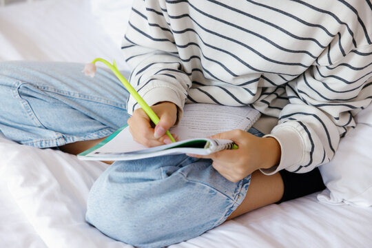 Partial of female school student writing in notebook on bed at home