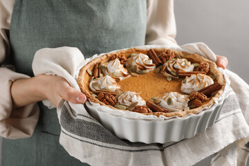 Woman holding homemade pumpkin pie with whipped cream, seeds and cinnamon on grey background, closeup