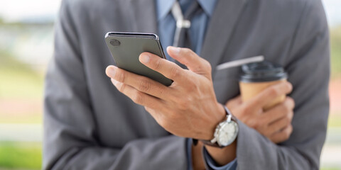 Close up of a young Asian businessman using a smartphone and holding a coffee cup in a professional outdoor setting.
