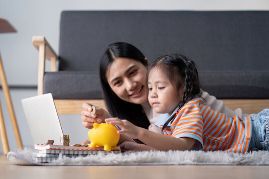 Diverse mother and daughter engaging in saving money together with a yellow piggy bank and laptop at home, enhancing financial literacy.