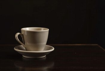Obraz premium A solitary beige coffee cup rests on a matching saucer atop a dark brown wooden table against a stark black backdrop. Simple, elegant, and minimalist.