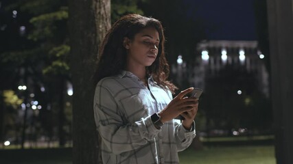 A young African American woman walks alone down a quiet city street in the evening. She is focused on her cellphone, surrounded by soft night lights. - Powered by Adobe