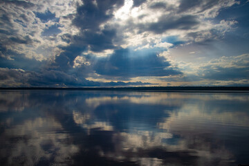Dramatic cloudy sky with sun rays over a calm river, reflecting clouds.
