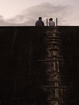 Two young men sit at the top of a high wall with makeshift ladder.