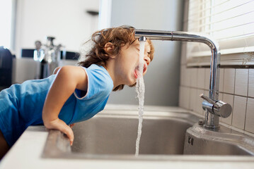 Young boy drinks water directly from kitchen sink at home