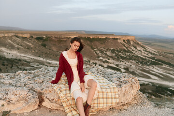 Woman relaxing on rock with plaid blanket in front of majestic mountain, leisure travel adventure lifestyle concept