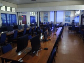 Fototapeta premium Blurred image of a modern office setting, featuring rows of workstations with computers on wooden desks. Blue office chairs align the desks, and the walls are adorned with windows and posters. The lig