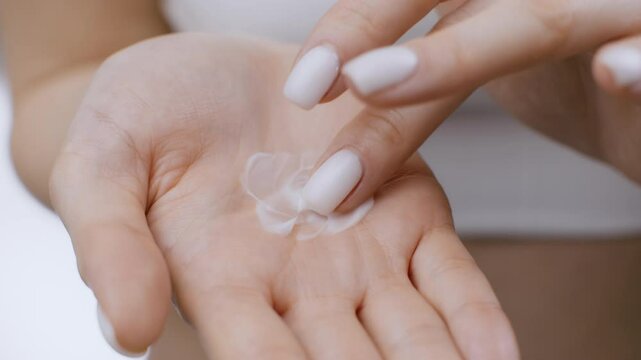 A young Caucasian woman shows a small amount of skin care cream in her hand, preparing to pamper herself. She stands against a clean, bright background focused on body care.
