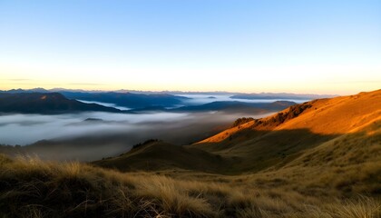 Sunrise over misty mountain range with golden grassy slopes, blue sky, and distant peaks fading into the horizon.