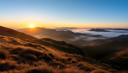 Sunrise over misty mountain range with golden grassy slopes, blue sky, and distant peaks fading into the horizon.