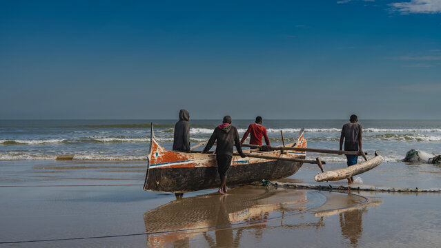 A group of people are carrying a pirogue  to the ocean. An old worn wooden fishing boat. The waves are foaming, spreading across the beach. Reflection on wet, smooth sand. Blue sky, clouds. Madagascar