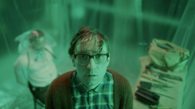 Mad serial killer with wet hair and large glasses standing in plastic-covered room with green lighting, looking into camera with excessive blinking, tied victim in background. Video portrait