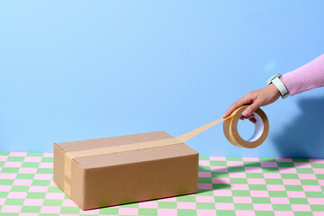 Hand Sealing Cardboard Box with Adhesive Tape on Checkered Table