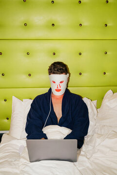 Male Wearing Red Light Mask Using Laptop in Bed