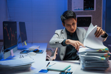 Asian businessman searching through documents at night in office with computer showing stock market...