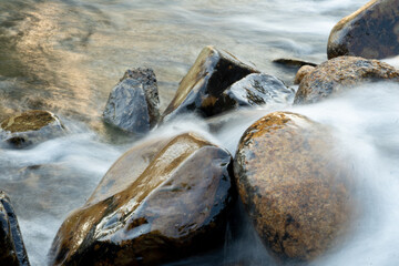 Shows a close-up of a riverbed with smooth  rounded stones. Water is flowing gently over the rocks creating a blurred and ethereal effect.