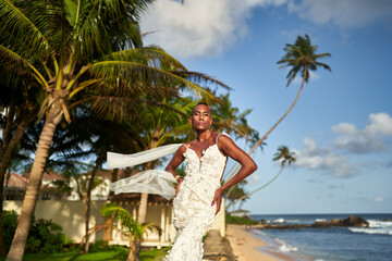 Person in white bridal dress on tropical beach. Ambiguous gender fluid black model poses between palm trees, ocean in background. LGBTQ-friendly wedding concept. Destination bridal fashion.