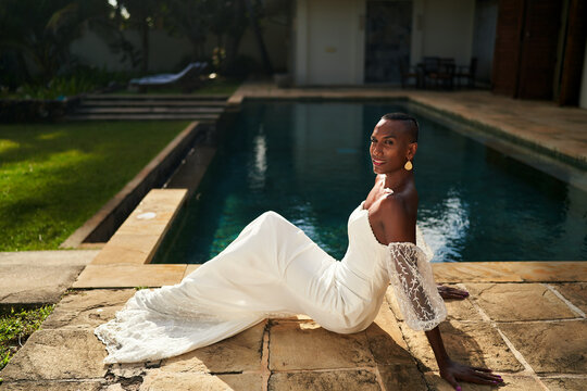 Black person in stylish white bridal dress poses by villa pool. Gender fluid fashion trend. LGBTQ friendly setting for chic wedding or event. Ethnic diversity. Summer day vibe. - Powered by Adobe