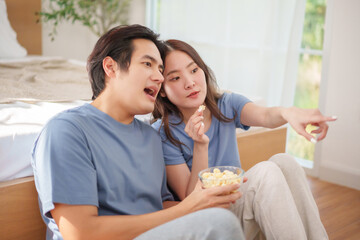 Asian couple sits together on the floor, happily sharing popcorn and engaging in animated conversation while enjoying their time at home. Watching television together
