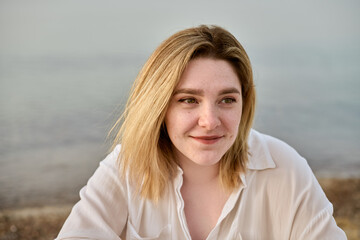 Young woman with blonde hair appears pensive by beach, evoking tranquility and reflection. Soft lighting and serene beach setting amplify sense of calm and introspection