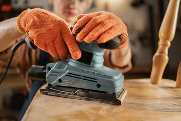 female carpenter polishing old chair restoring furniture in workshop using sanding machine