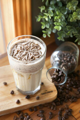 A creamy iced coffee beverage with an intricate foam design is presented on a wooden board alongside scattered coffee beans and an overturned glass jar, with lush greenery in the background