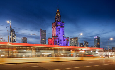 Warsaw Palace of Culture glows in red, white, and blue against the night sky, surrounded by modern skyscrapers. Light trails from passing cars and trams create a dynamic sense of motion  © elena_suvorova