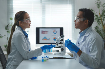 Two scientists wearing lab coats and protective eyewear discuss research findings in a laboratory. A microscope and data charts on a screen highlight their scientific work environment