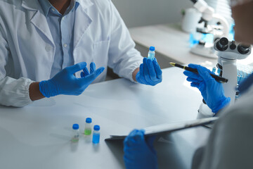 Two scientists wearing lab coats and gloves are examining a vial containing blue liquid, discussing research findings and analyzing data in a laboratory setting with a microscope nearby