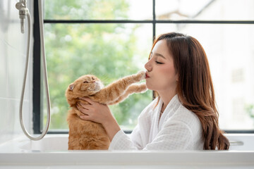 Asian young woman sitting in bathtub with her domestic cat in bathroom. 