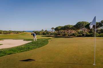 Golfer playing near a sand bunker on a scenic golf course with a flag, lush greenery, and ocean in the background under a sunny blue sky.
