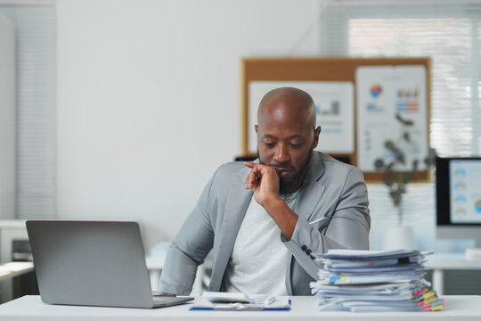 Exhausted african american businessman sitting at desk with hand on chin, looking stressed and overwhelmed by paperwork and workload pressure in office
