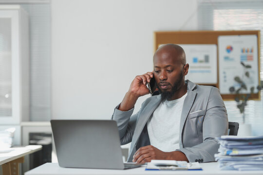 Serious african american businessman wearing gray jacket talking on cell phone and using laptop while analyzing documents at workplace in modern office
