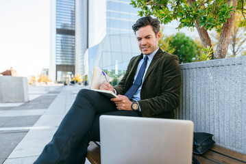 Businessman writing notes in notebook while working remotely outdoors
