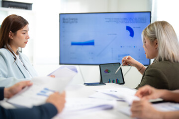 Fototapeta premium Two professionals analyze data during a meeting, with charts displayed on a screen, surrounded by documents and engaged in discussion.
