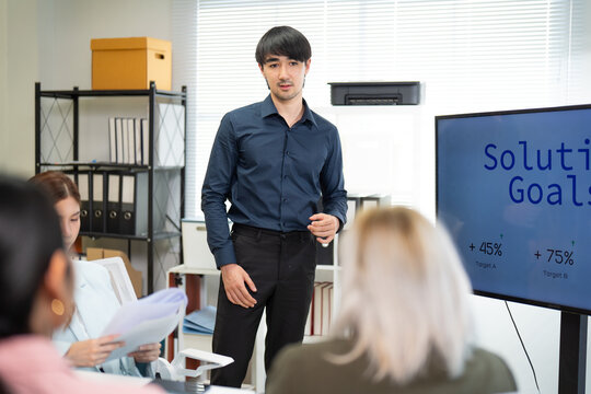 A young professional presents solutions and goals in a meeting, engaging with colleagues while showcasing a digital display.